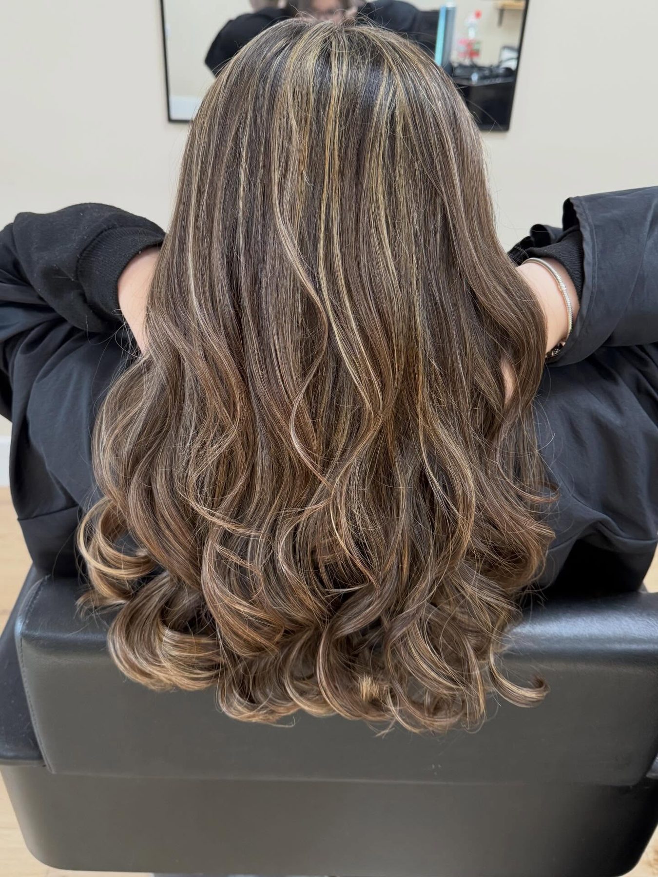 Long curled hair with soft highlights, seen from behind in the salon chair
